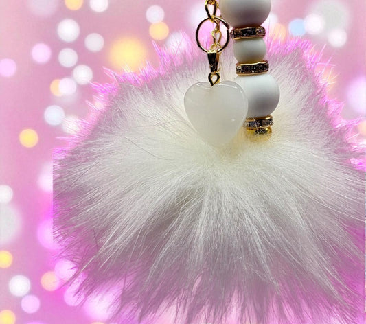 White fluffy pom-pom keychain with heart-shaped beads on a pink blurred background