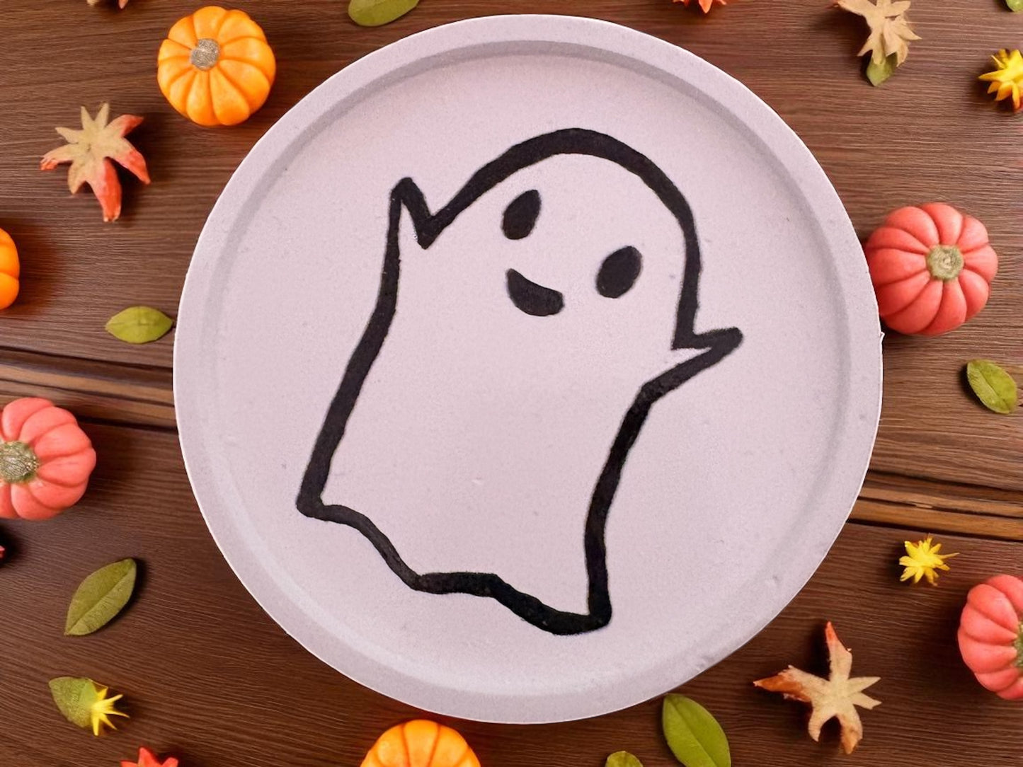 White plate with a black ghost drawing on a wooden surface with Halloween decorations.
