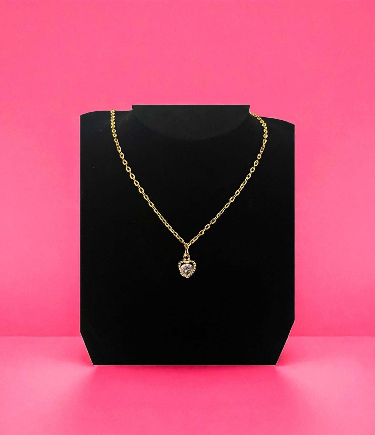 Gold necklace with a heart-shaped pendant on a black stand against a pink background