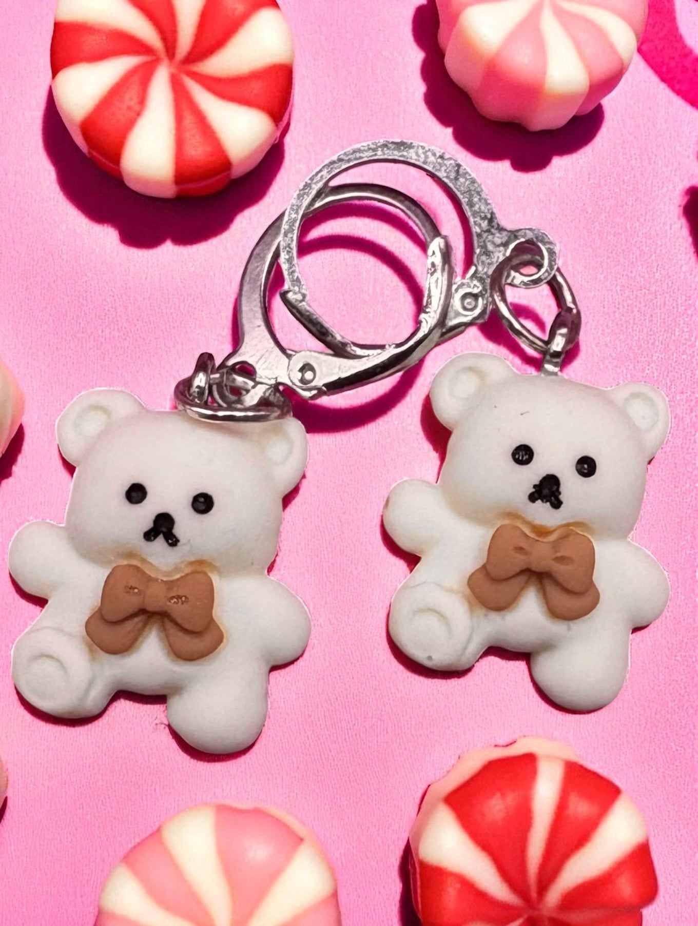 Two white teddy bear earrings with brown bow ties on a pink background with candy.