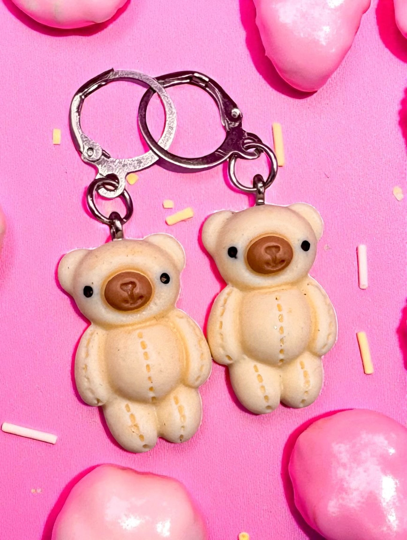 Two teddy bear earrings on a pink background with candy-like shapes.