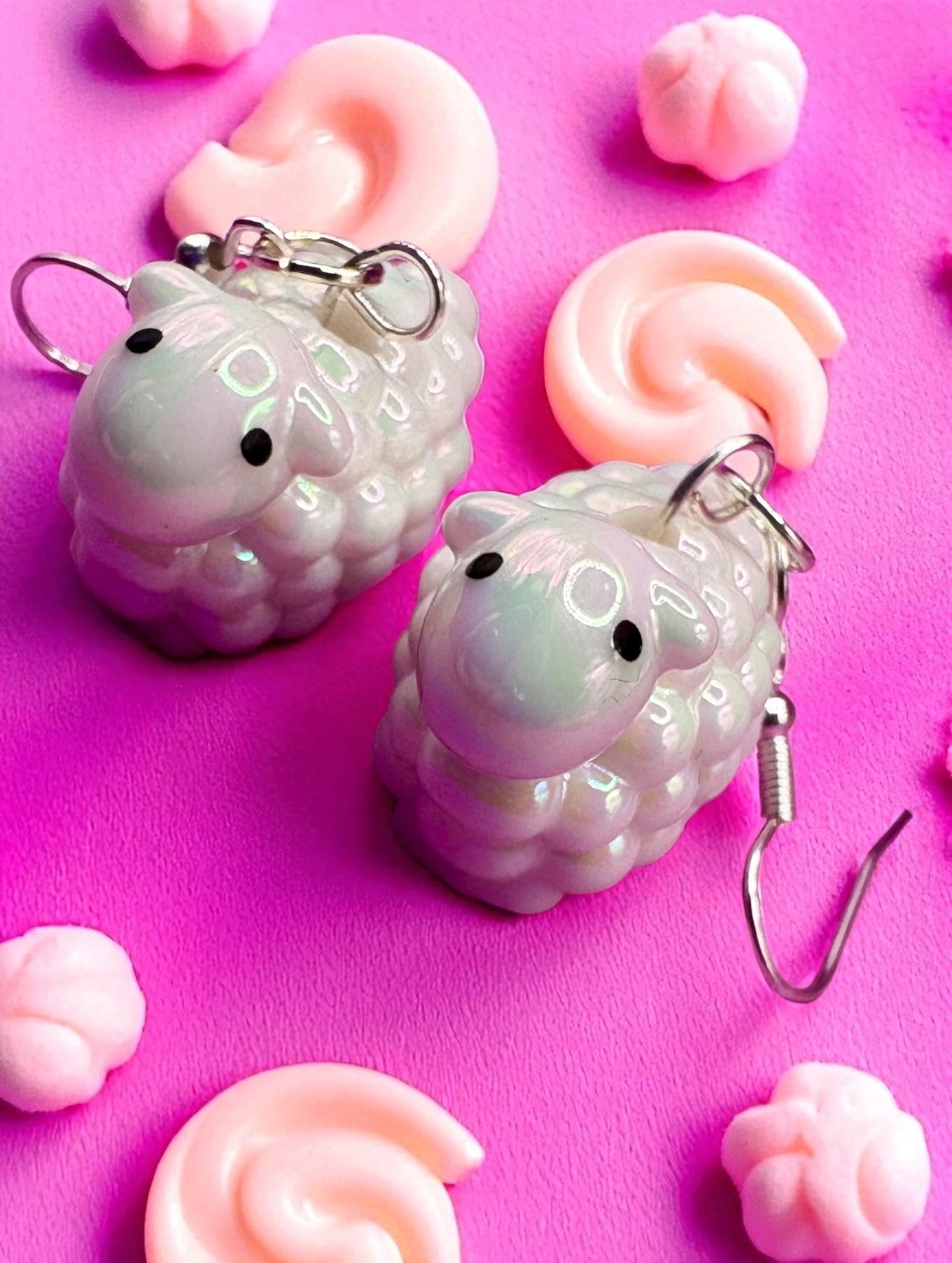Two sheep-shaped earrings on a pink background with pink sheep-shaped candies.