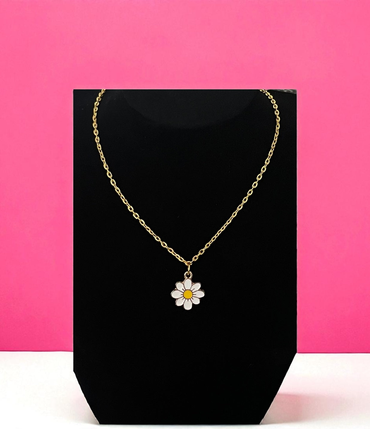 Gold necklace with a daisy pendant on a black stand against a pink background