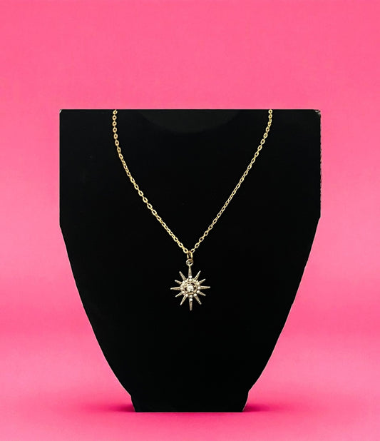 Gold necklace with a star-shaped pendant on a black stand against a pink background