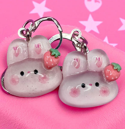 Two pink cat-shaped keychains with strawberry decorations on a pink background with star patterns.