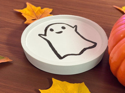 Round white coaster with a ghost design on a wooden surface with autumn leaves and a pumpkin.