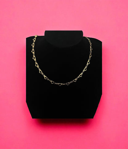 Gold heart-shaped chain necklace on a black stand against a pink background