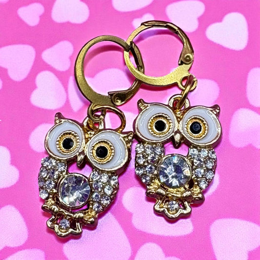 Gold owl-shaped earrings with gemstones on a pink background with heart patterns