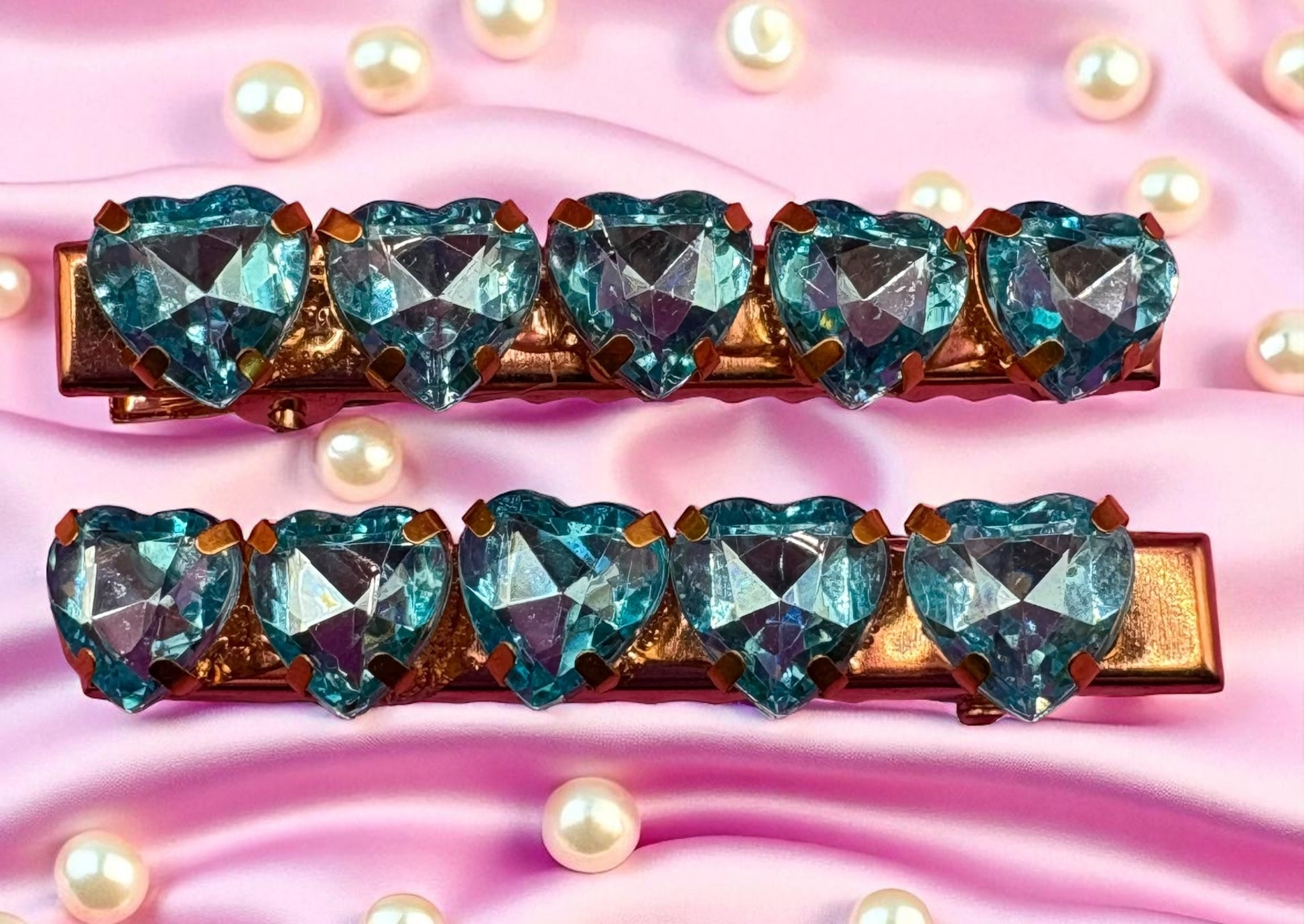 Two hair clips with heart-shaped blue gemstones on a pink background with pearls.