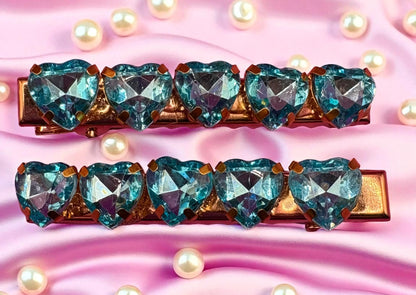 Two hair clips with heart-shaped blue gemstones on a pink background with pearls.