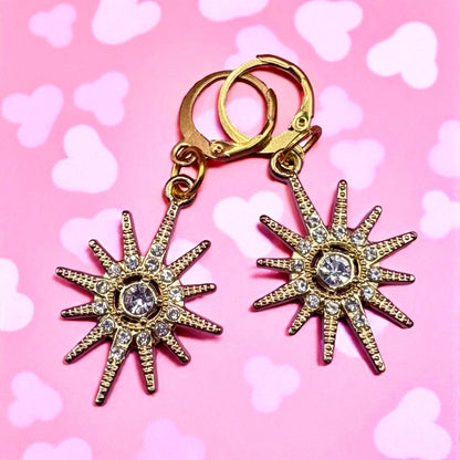 Gold star-shaped earrings with rhinestones on a pink background with heart shapes.