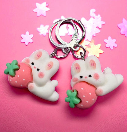 Two cute keychains shaped like bunnies holding strawberries on a pink background with star-shaped decorations.