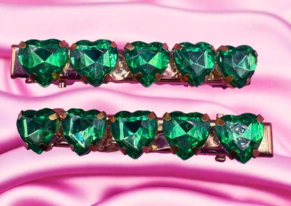 Two green heart-shaped gemstone hair clips on a pink fabric background