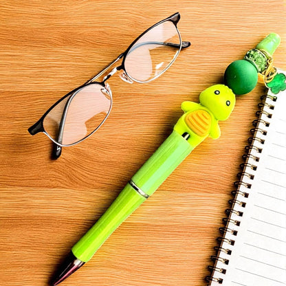 Green turtle-shaped pen on a wooden surface with glasses and a notebook.