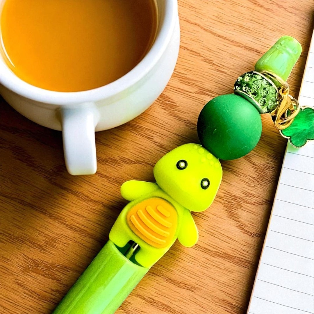 Green dinosaur-shaped pen on a wooden surface with a cup of tea and notebook.