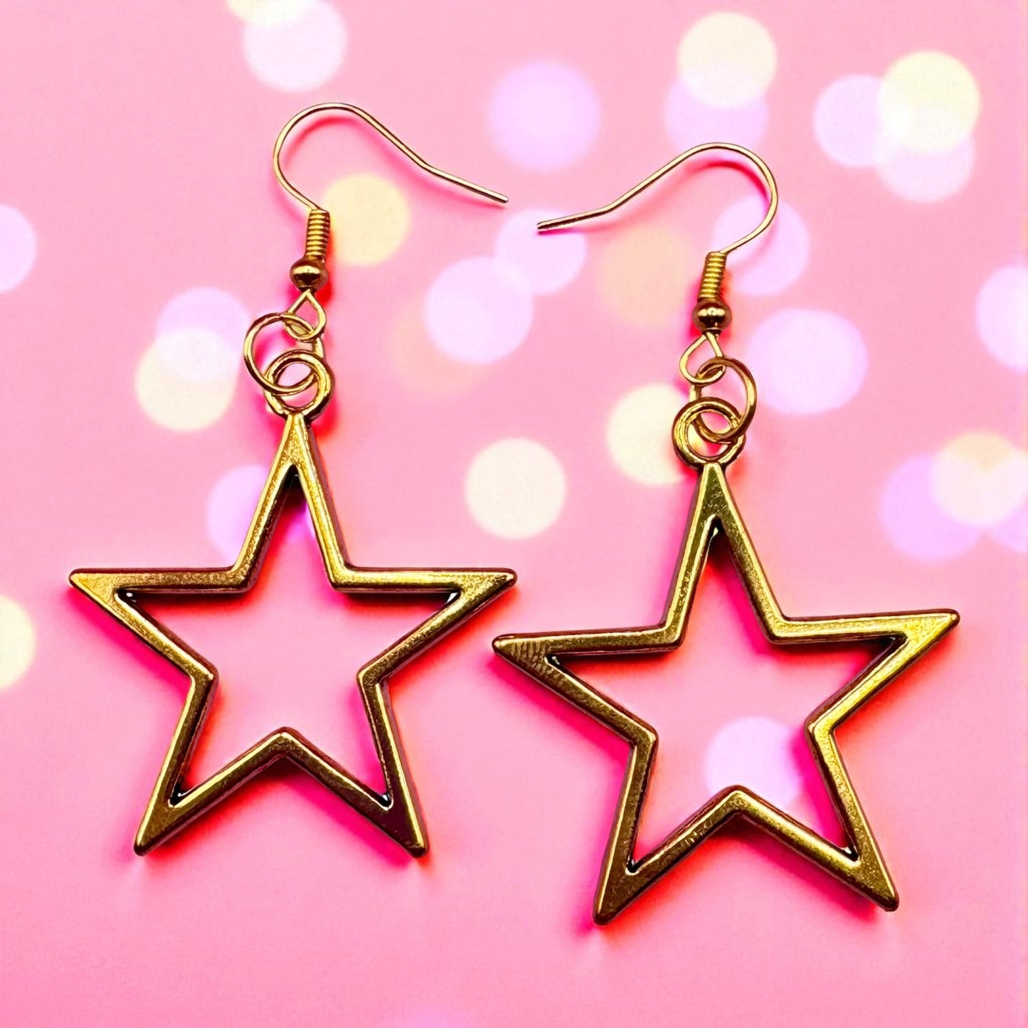 Gold star-shaped earrings on a pink background with bokeh lights