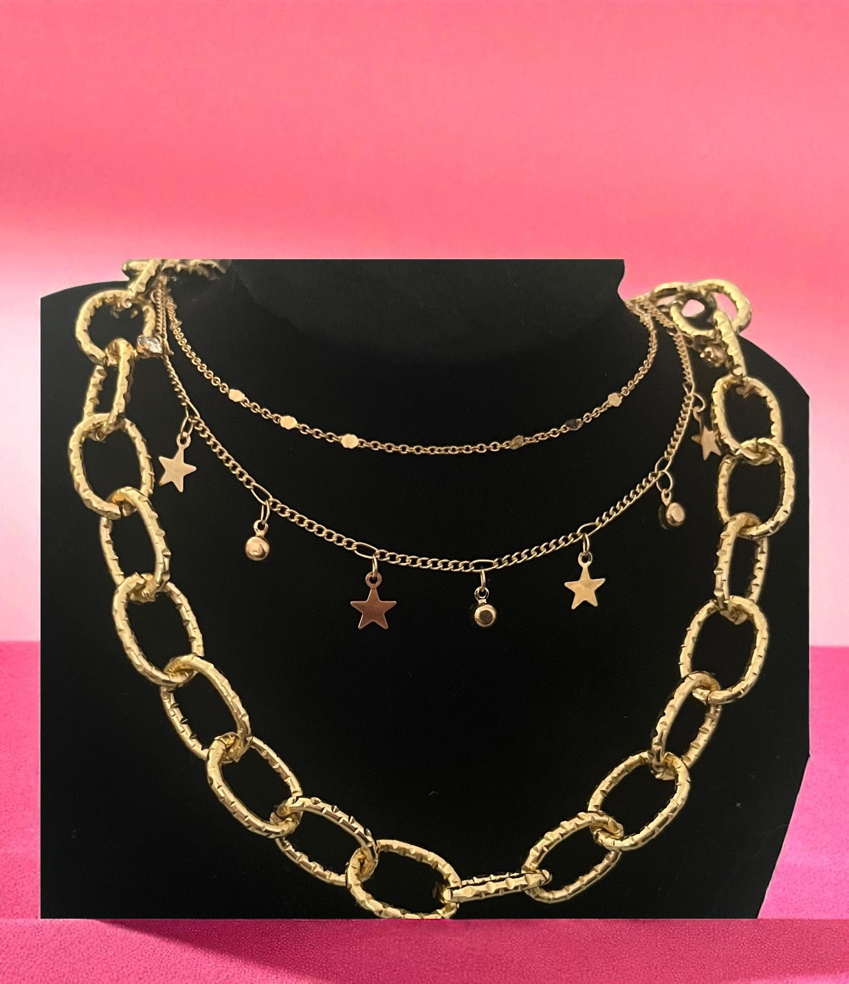 Gold chain necklace with star pendants on a black stand against a pink background