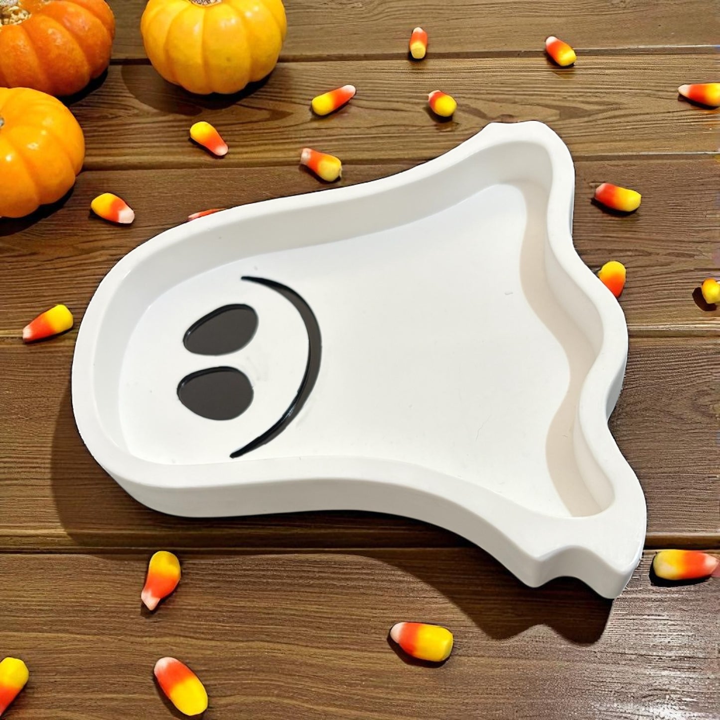 White ghost-shaped tray with black eyes on a wooden surface with pumpkins and candy corn.