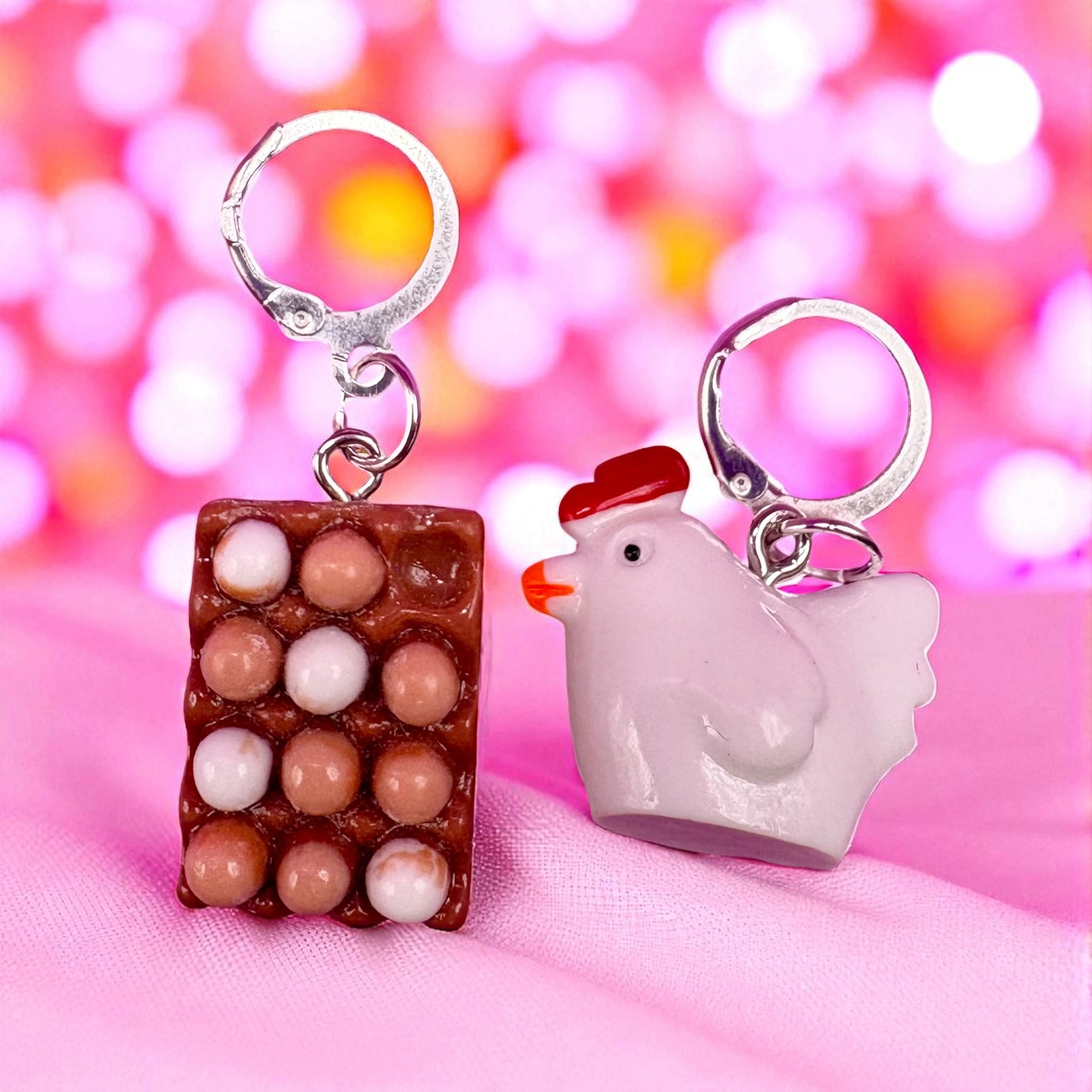 Two  earrings, one resembling a chocolate bar with white and brown spheres and the other a chicken, on a pink background.