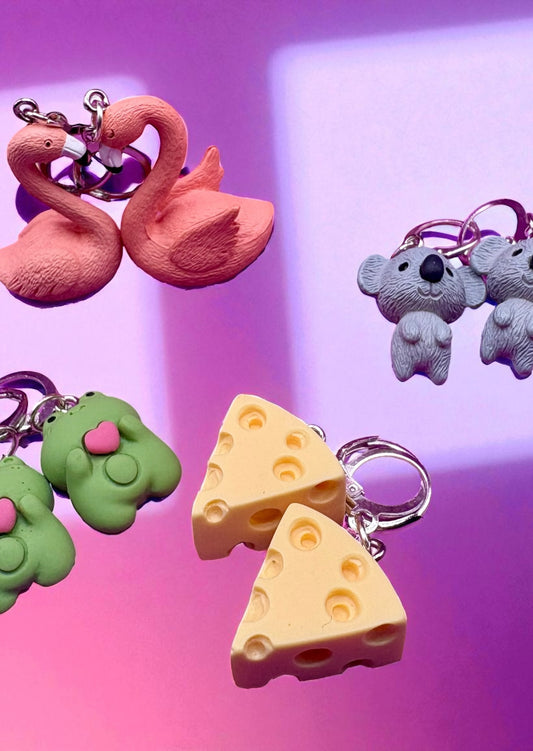 Colorful keychains including a pink swan, green creature, purple flower, and cheese slices on a gradient background.