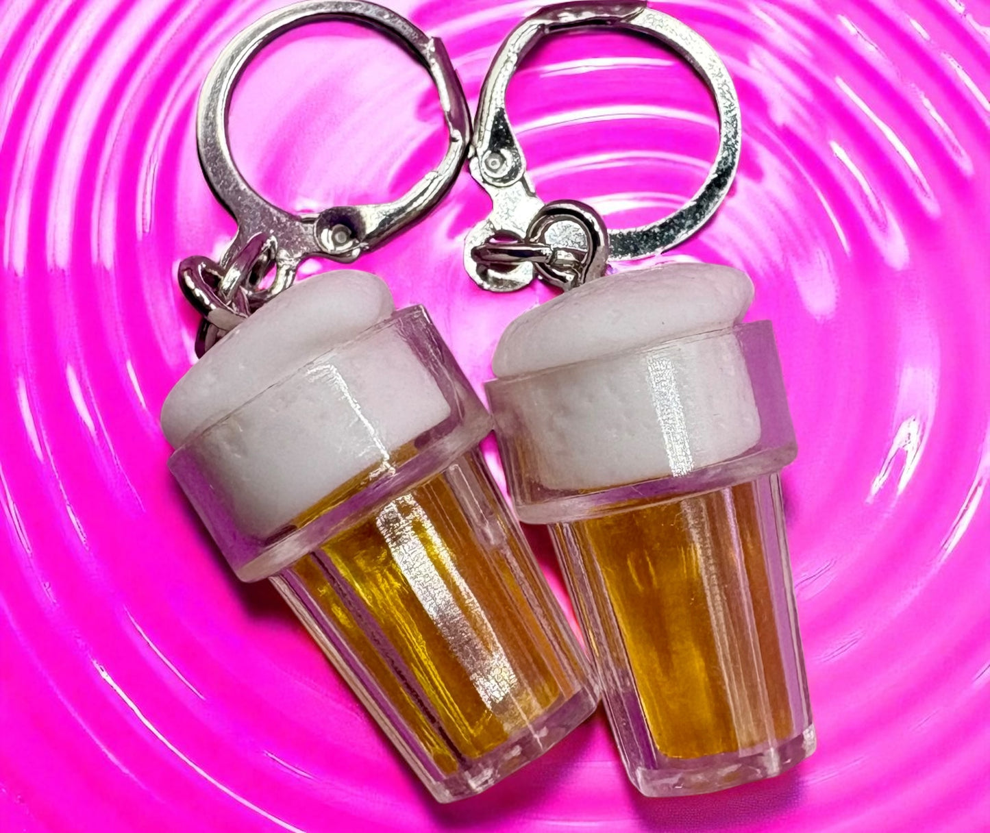 Two  earrings shaped like beer bottles with white caps on a pink swirling background