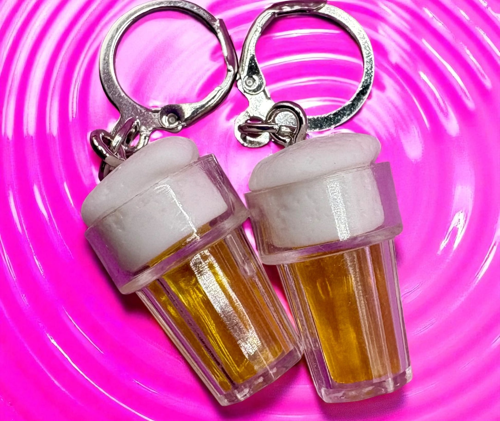 Two  earrings shaped like beer bottles with white caps on a pink swirling background