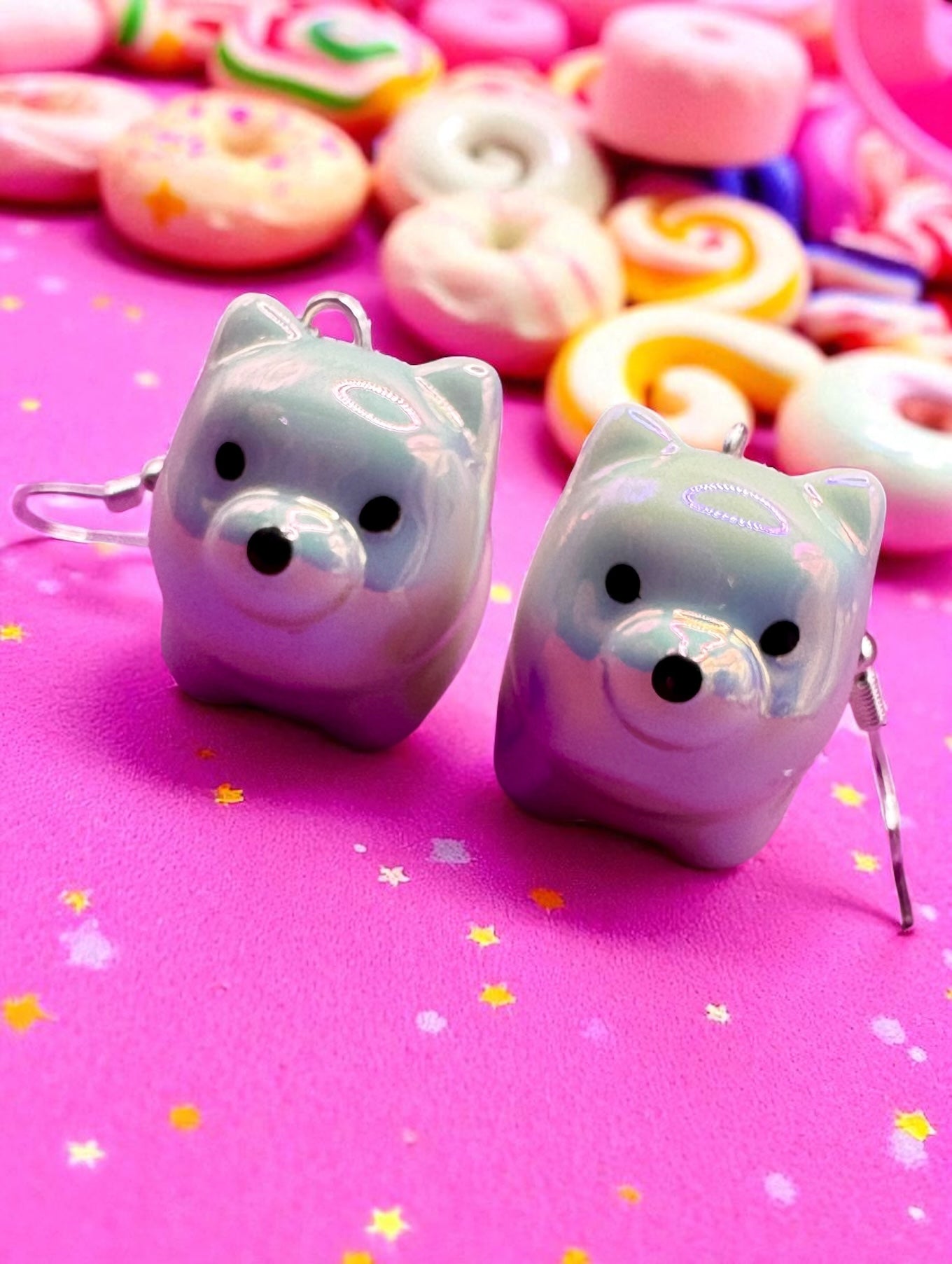 Pair of cat-shaped earrings on a pink background with colorful donuts and candies.