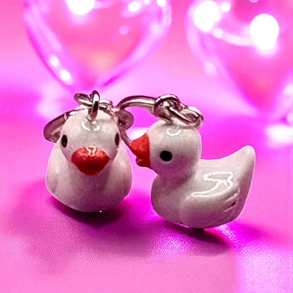 Pair of white duck-shaped earrings with red beaks on a pink background