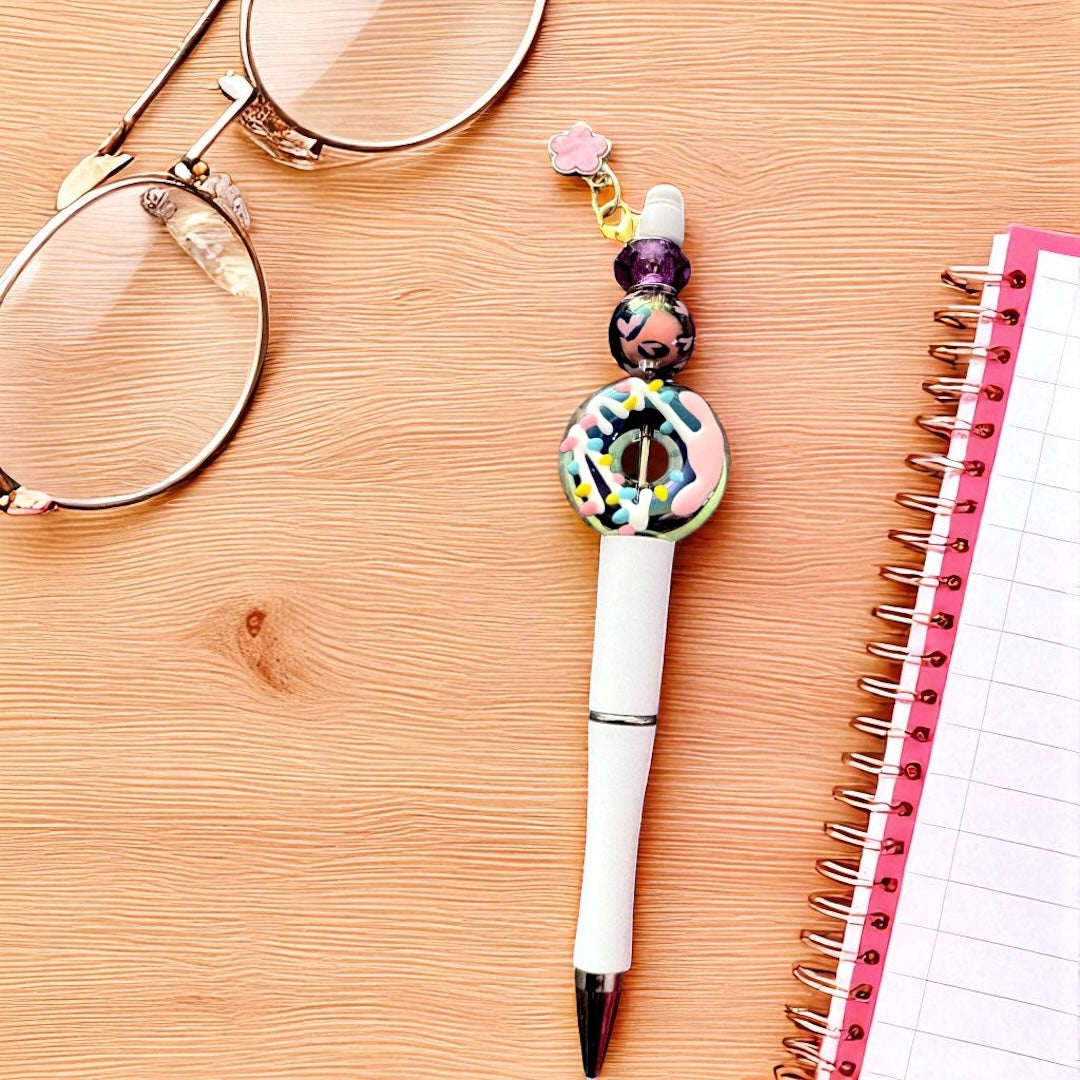 Decorative pen with beads on a wooden surface next to a notebook and glasses.
