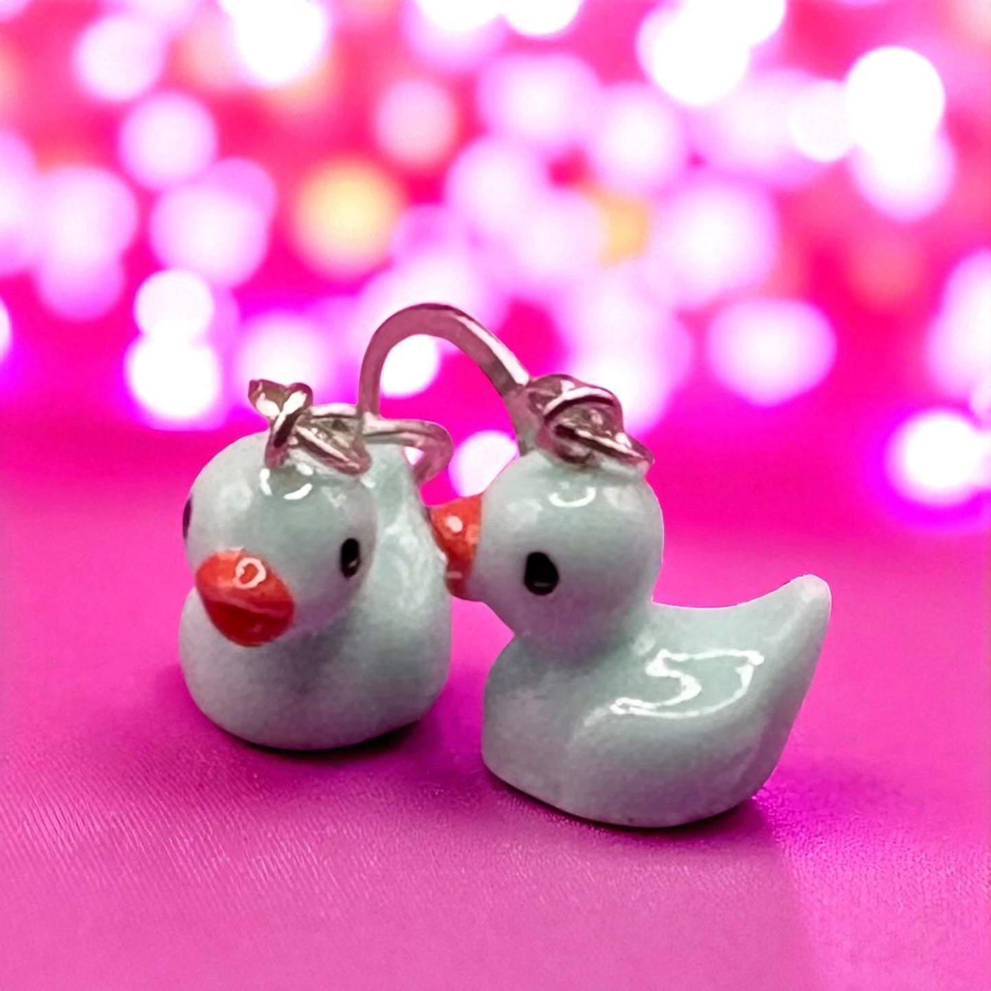 Pair of duck-shaped earrings on a pink background with bokeh effect