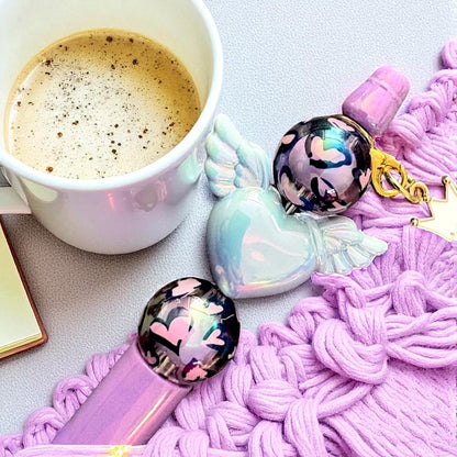 Pink pen with heart-shaped cap and angel wings, next to a cup of coffee on a textured surface.