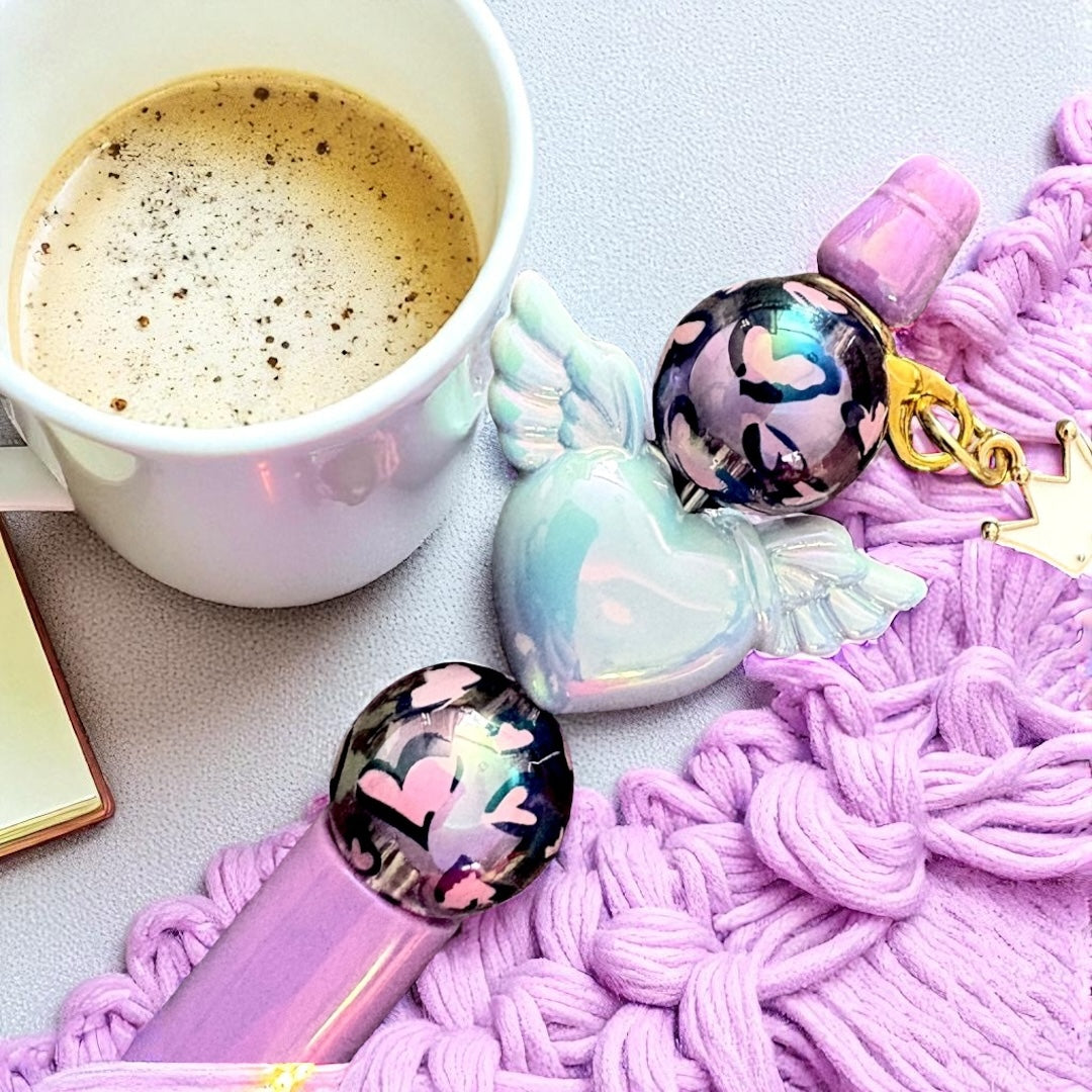 Pink pen with heart-shaped cap and angel wings, next to a cup of coffee on a textured surface.