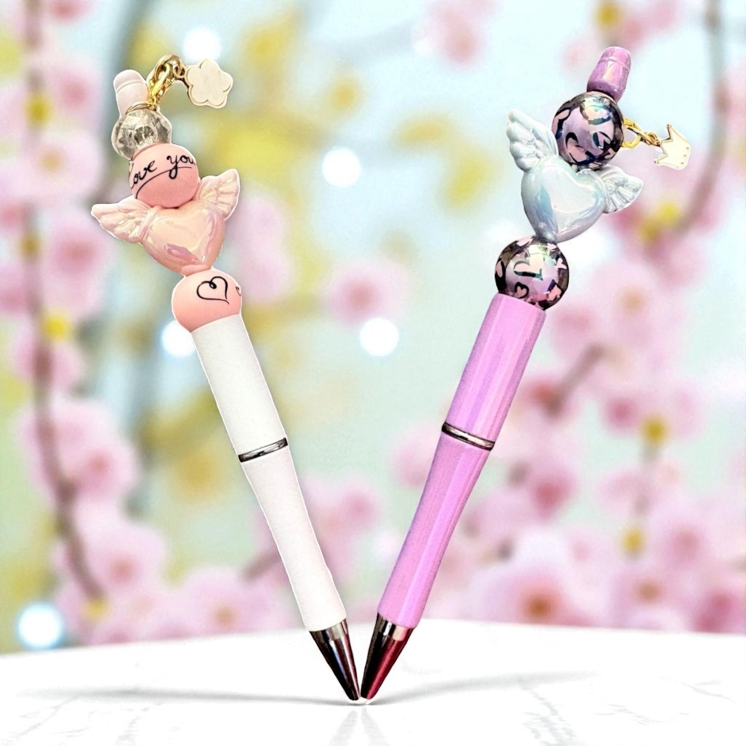 Two decorative pens with angel charms on a blurred floral background