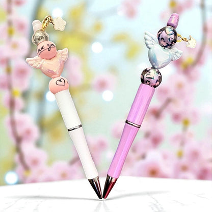 Two decorative pens with angel charms on a blurred floral background